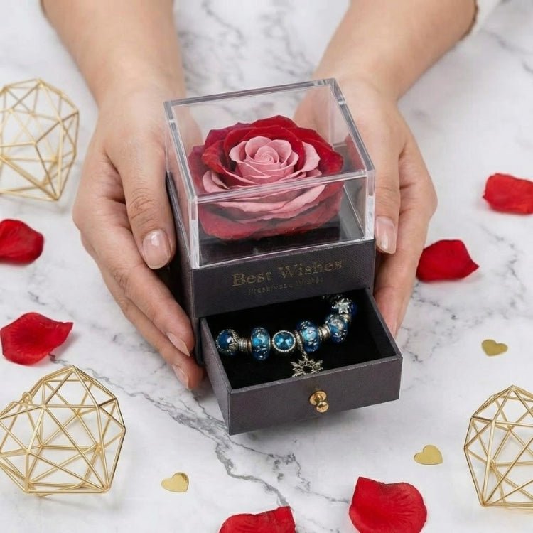 Hands presenting a romantic two-tone red and pink eternal rose gift box with a visible snowflake charm bracelet in the bottom drawer.