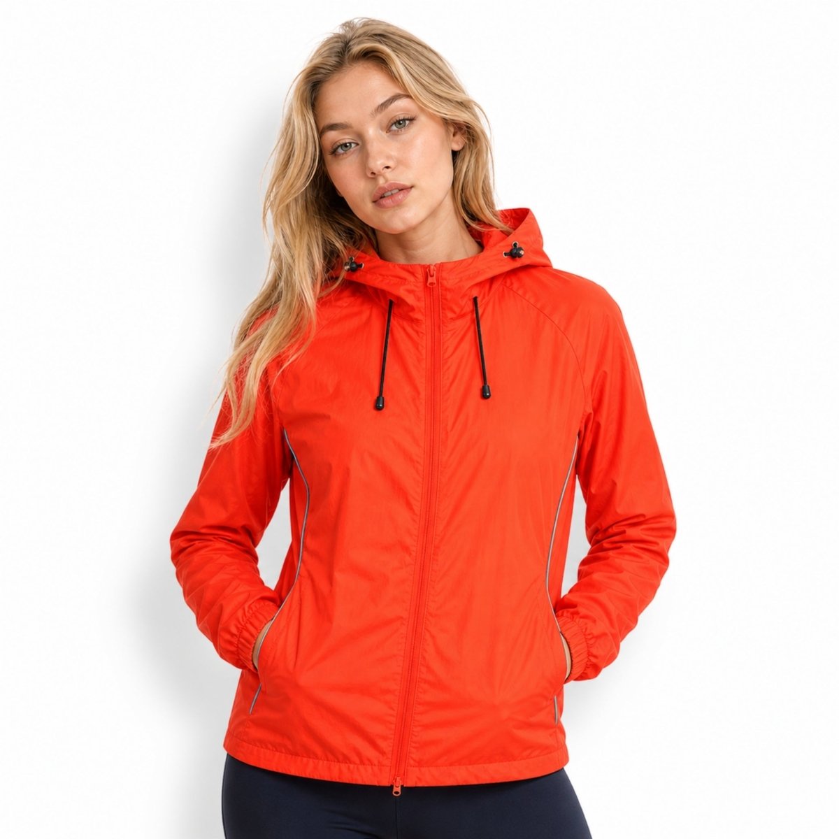 Blonde female model wearing Kouvr bright orange lightweight hooded windbreaker jacket with hands casually resting in front pockets.
