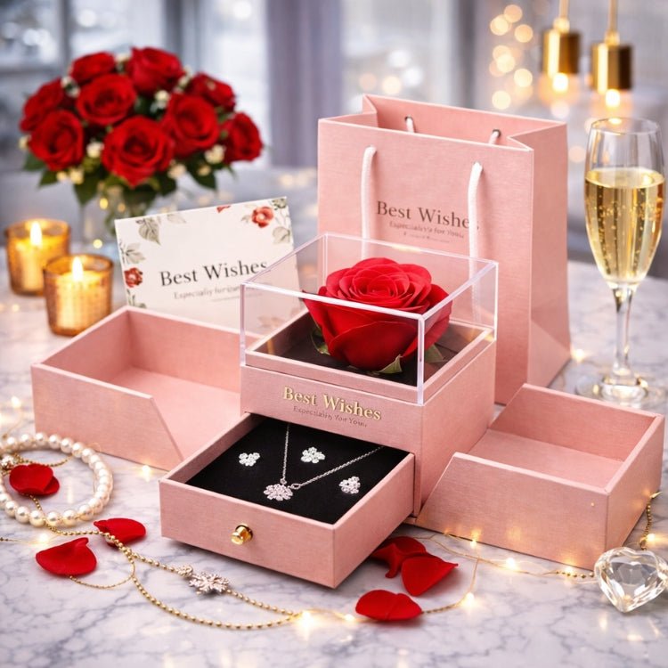 Open pink jewelry gift box featuring a preserved red rose under a clear lid and a drawer containing a silver necklace and earrings, set against a romantic marble background with candles and champagne